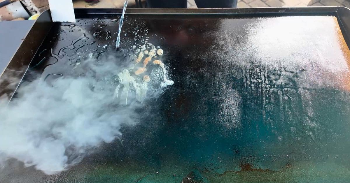 Hot water being poured onto a Blackstone griddle, creating steam as it loosens stuck-on food residue for cleaning.
