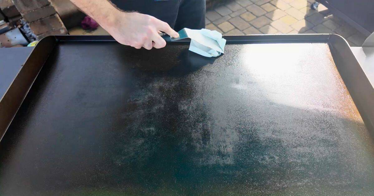 A person using a metal spatula and a paper towel to wipe down a clean, well-seasoned Blackstone griddle after cooking.