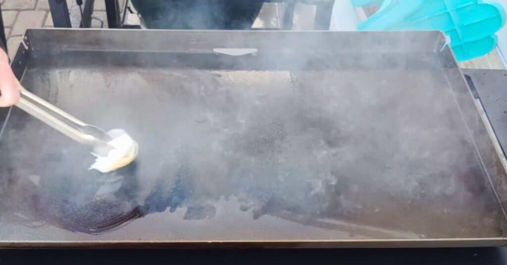 a blackstone griddle smoking from oil being burnt off.  metal tongs and a paper towel moving the oil around.