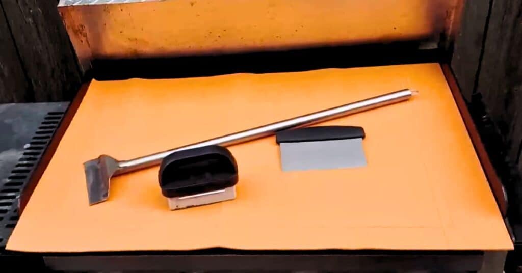 An orange silicone mat on a blackstone griddle with a scrapper and pumice stone on top.