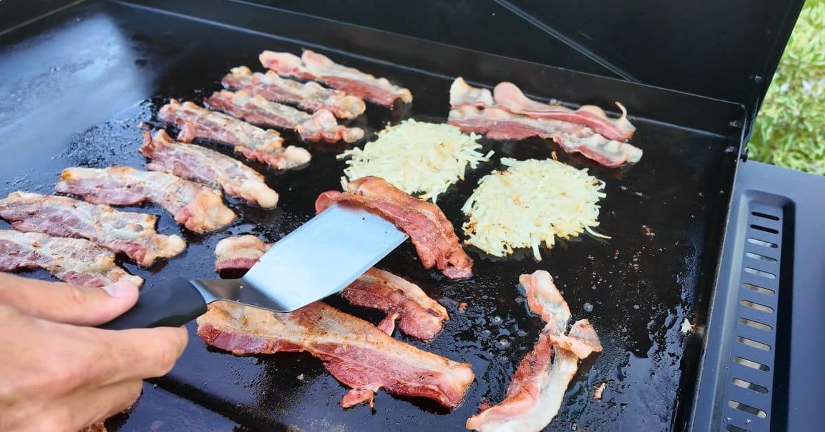 Close-up of bacon and hash browns cooking on a Blackstone griddle, with a hand using a spatula to flip crispy bacon