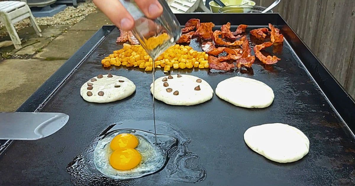 Cooking a full breakfast on a Blackstone griddle with chocolate chip pancakes, crispy bacon, diced potatoes, and eggs cracked onto the hot surface