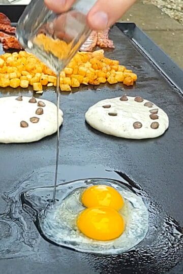 2 whole eggs being poored on a griddle with pancakes bacon and potatoes in the background.