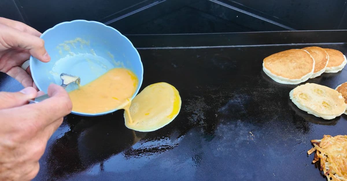Scrambeled eggs in a blue bowl being poured on a griddle next to banana pancakes