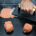 ChatGPT said: A hand presses down a ball of raw ground beef on a griddle using a cast iron burger press with a sheet of parchment paper underneath, flattening it into a smash burger patty. Two more unpressed beef portions are on the hot, oiled griddle nearby. Steam rises as the meat sears.