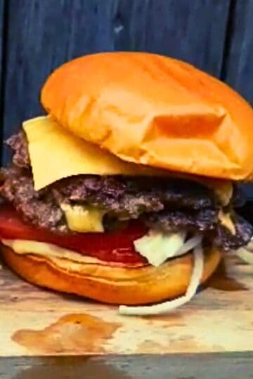 A freshly cooked smash burger stacked on a wooden board, featuring a shiny toasted bun, two crispy beef patties, melted American cheese, sliced tomato, shredded lettuce, onions, and a layer of sauce. The burger is slightly messy and oozing with flavor.