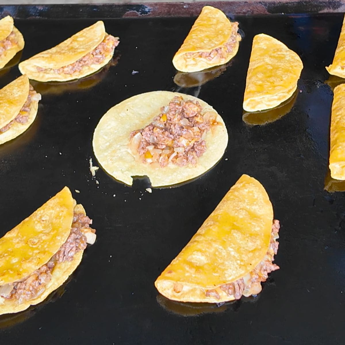 Corn tortillas filled with a seasoned ground beef mixture being cooked and folded into tacos dorados on a Blackstone griddle.