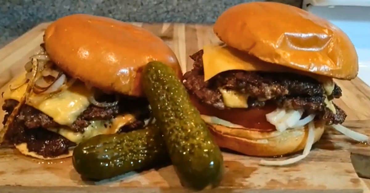 Two smash burgers cooked on a blackstone griddle with two pickles in front.