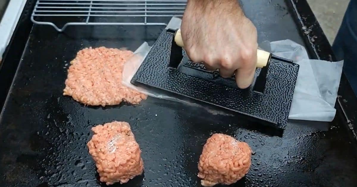 Burgers added to the griddle and being smashed down by a hand and burger tool.