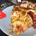 A grilled chicken breast topped with crispy bacon, caramelized onions, and shredded Colby Jack cheese, served on a white plate. A red arrow points to the dish, with a flat-top griddle and additional servings visible in the background.