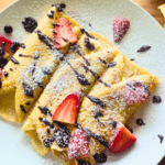 A plate of golden-brown crepes folded and filled with cream cheese, topped with fresh strawberry slices, drizzled with chocolate syrup, and dusted with powdered sugar. The plate rests on a wooden cutting board, with an additional rolled crepe visible in the background.