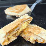 A golden-brown breakfast crunchwrap cut in half, revealing layers of crispy hash browns, scrambled eggs, bacon, and melted cheese, sitting on a Blackstone griddle. Another whole crunchwrap is in the background with a spatula resting nearby.