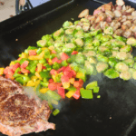 A sizzling steak, diced bell peppers, Brussels sprouts, and crispy roasted potatoes cooking on a Blackstone griddle.