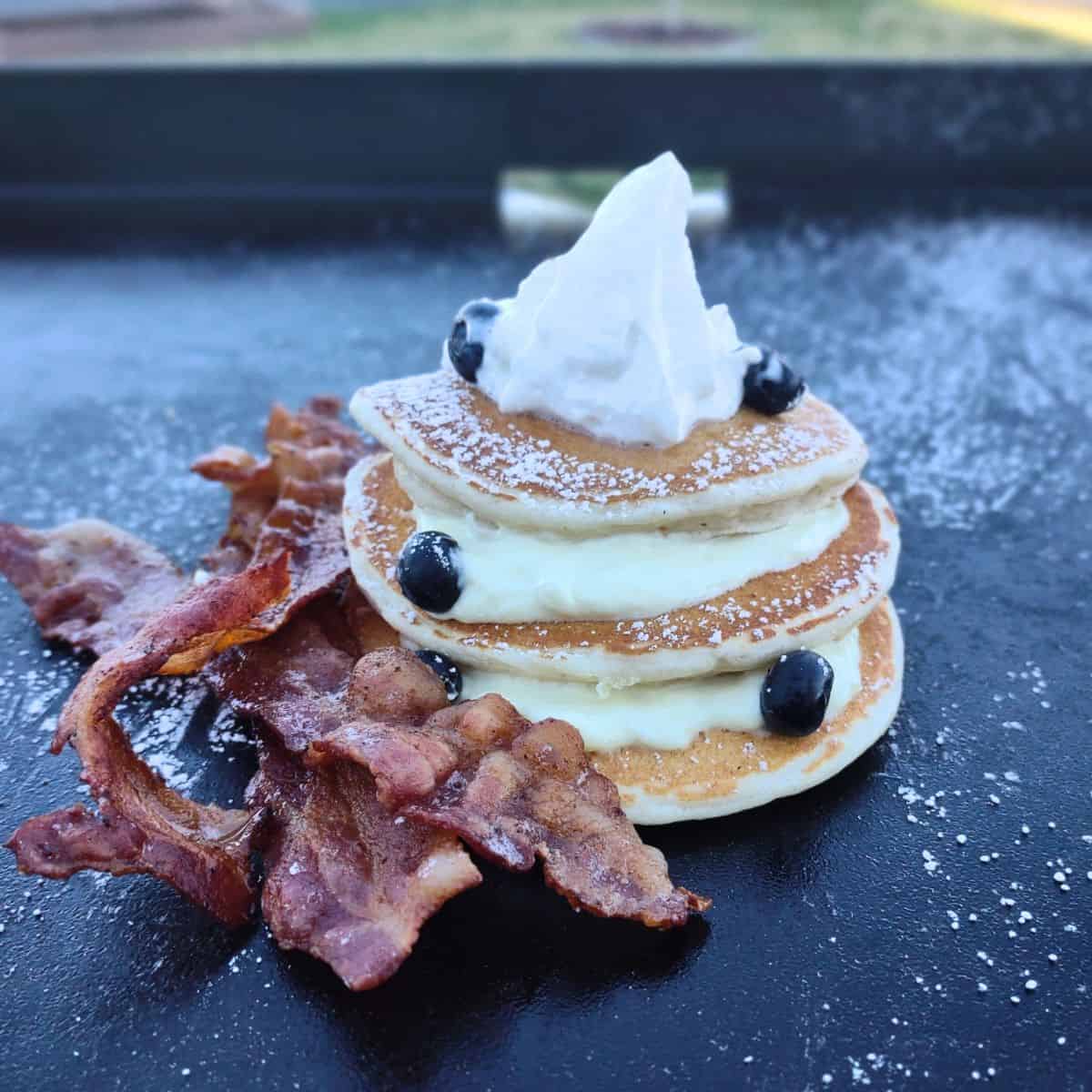 Blueberry pancakes stacked on a blackstone griddle with a c cream cheese filling. Whipped cream on top and bacon on the side.