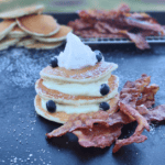 Stack of blueberry pancakes with whipped cream and powdered sugar on a Blackstone griddle, served with crispy bacon. More pancakes and bacon rest in the background on a cooling rack.