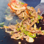 Steak, red bell pepper, yellow bell pepper, white onion tossed on a blackstone griddle with two spatulas.