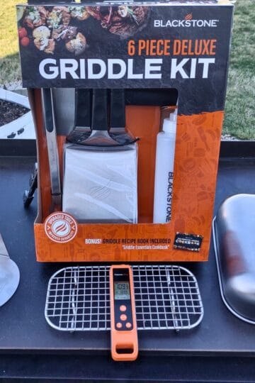 A Blackstone 6-piece deluxe griddle kit is displayed on a flat-top griddle outdoors. The kit includes two spatulas, a scraper, tongs, and a squeeze bottle. Also on the griddle are a burger press, a digital thermometer, a wire cooling rack, and a stainless steel basting dome. The background shows grass and a raised garden bed.