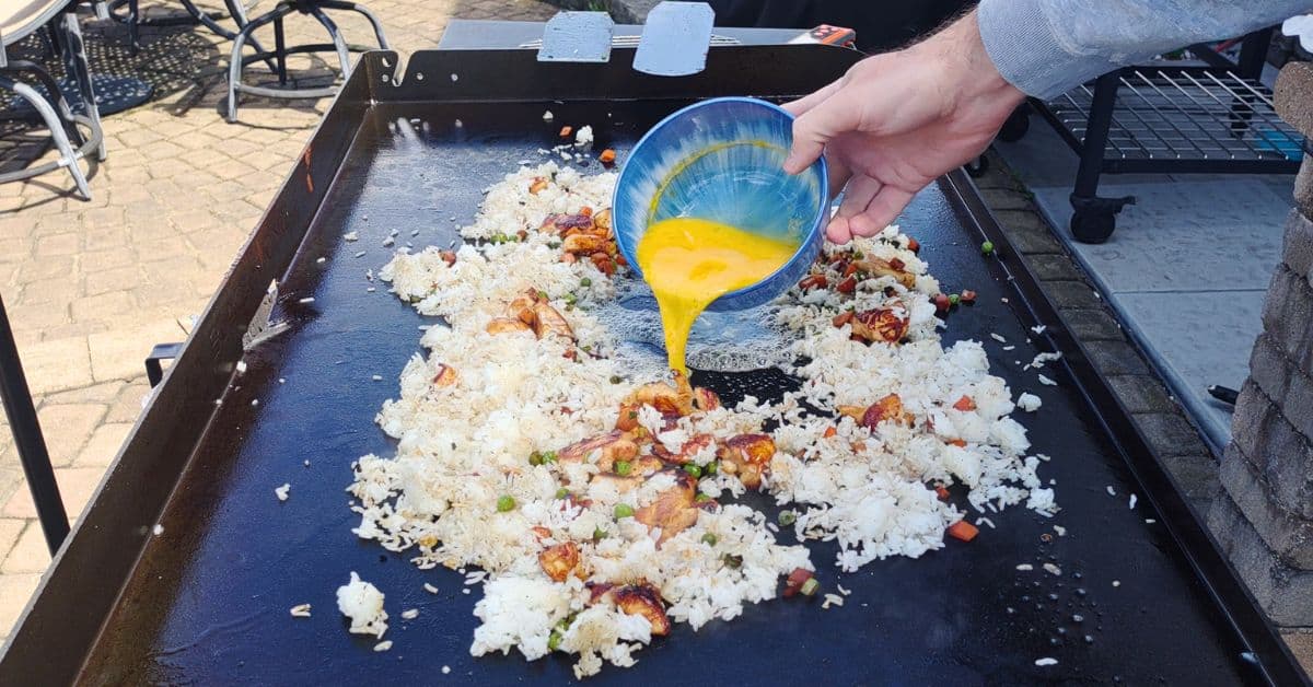 white rice and peas and onions cooking on a blackstone griddle.  a blue bowl pouring scrambled eggs in the rice.