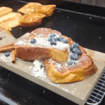 Thick slices of golden-brown challah French toast on a wooden board, topped with whipped cream, fresh blueberries, powdered sugar, and two cinnamon sticks, with more slices cooking in the background on a griddle.