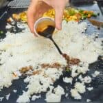 A hand pours dark soy sauce from a small plastic cup onto a bed of white rice cooking on a Blackstone griddle. In the background, mixed vegetables and scrambled eggs are also being cooked, suggesting the preparation of chicken fried rice.