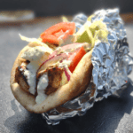 A freshly made chicken gyro wrapped in foil, resting on a griddle. The gyro is filled with grilled chicken pieces, sliced red onions, lettuce, tomatoes, and drizzled with a creamy white sauce, all wrapped in warm pita bread.