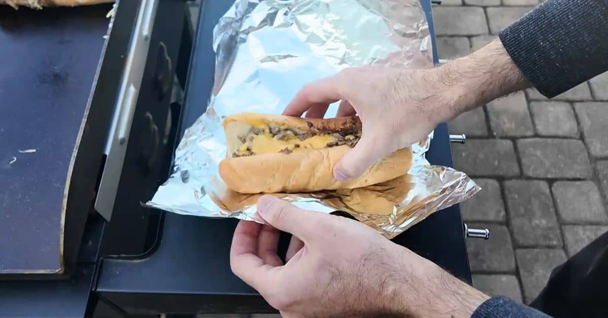 Close-up of hands wrapping a cheesesteak loaded with steak and melted cheese in foil, next to a griddle.