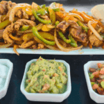 Grilled chicken fajitas served on a rectangular white platter with sautéed onions and bell peppers, fresh lime wedges, and warm tortillas. Accompanied by small dishes of sour cream, guacamole, and pico de gallo, all presented on a Blackstone griddle.