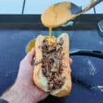 "Close-up of a hand holding a cheesesteak sandwich on a griddle, with melted Cheez Whiz being drizzled over thinly sliced ribeye steak and onions inside a soft hoagie roll."