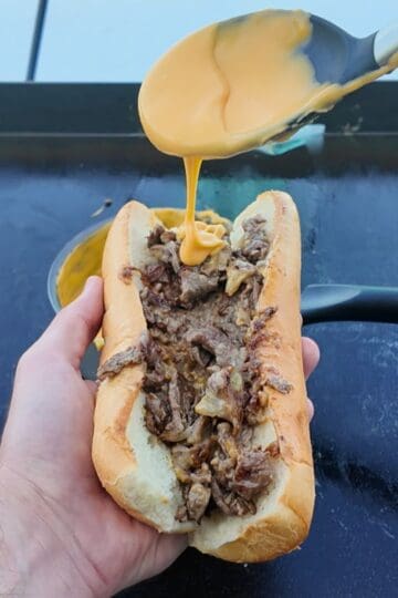 "Close-up of a hand holding a cheesesteak sandwich on a griddle, with melted Cheez Whiz being drizzled over thinly sliced ribeye steak and onions inside a soft hoagie roll."