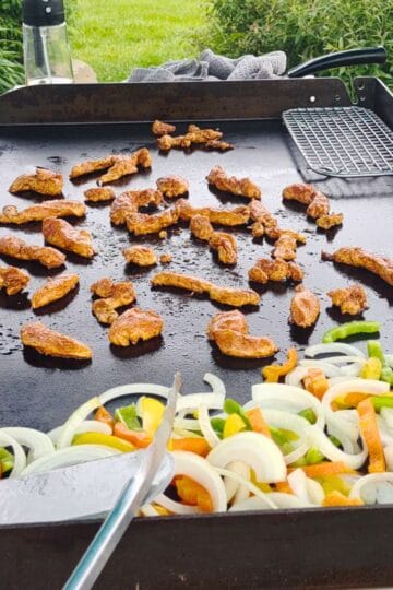 Chicken fajitas sizzling on a Blackstone griddle with steam rising, surrounded by colorful sliced onions and bell peppers. A hand is using a metal spatula to cook the mixture outdoors.
