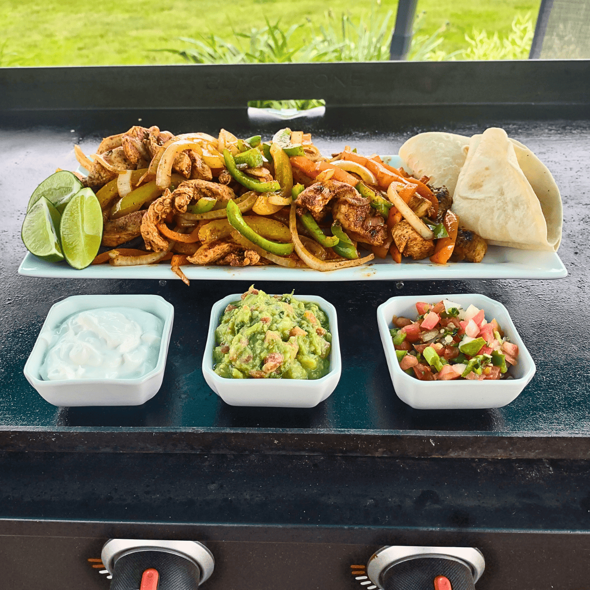 Grilled chicken fajitas served on a rectangular white platter with sautéed onions and bell peppers, fresh lime wedges, and warm tortillas. Accompanied by small dishes of sour cream, guacamole, and pico de gallo, all presented on a Blackstone griddle.