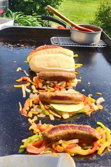 Italian sausages cooking on a Blackstone griddle with sautéed peppers and onions. Some sandwiches are assembled with melted cheese and hoagie rolls, while a small pot of red sauce warms on a wire rack in the background. The scene is outdoors on a sunny day with green grass and garden plants nearby.