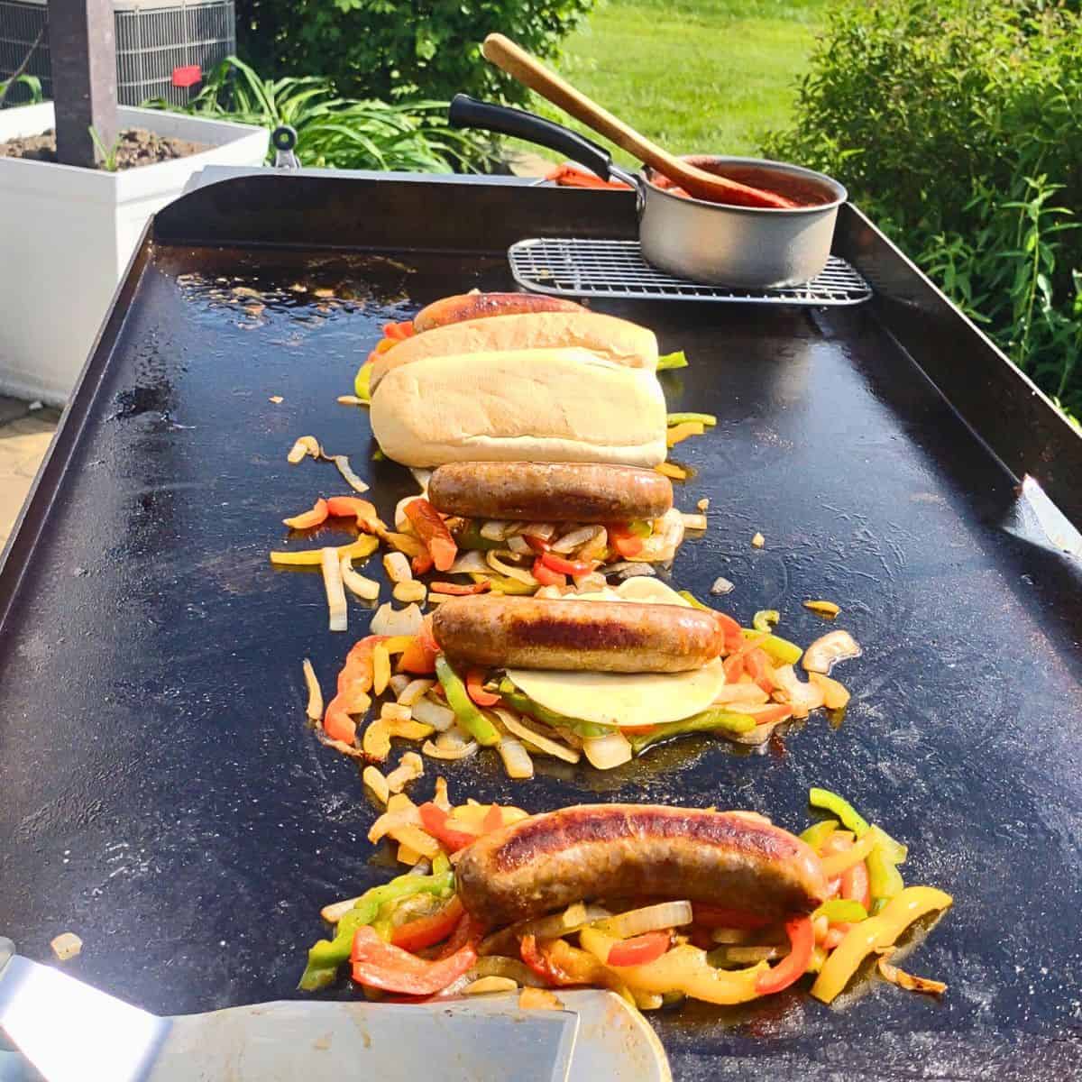 Italian sausages cooking on a Blackstone griddle with sautéed peppers and onions. Some sandwiches are assembled with melted cheese and hoagie rolls, while a small pot of red sauce warms on a wire rack in the background. The scene is outdoors on a sunny day with green grass and garden plants nearby.