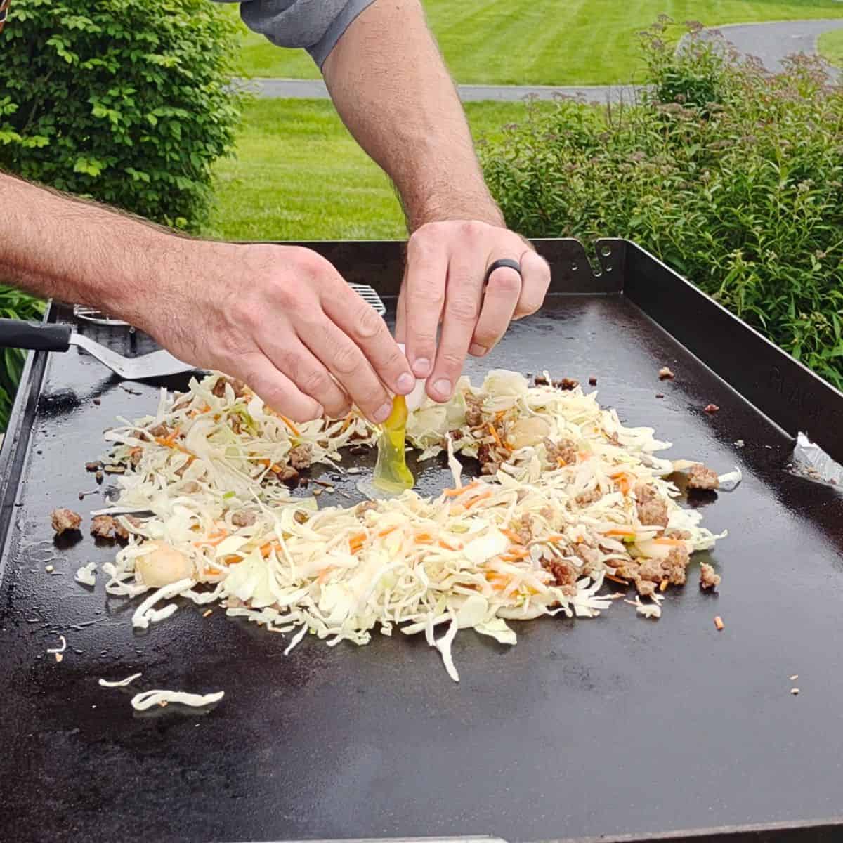 Egg roll in a bowl cooking on a Blackstone griddle with an egg cracked into the center of cabbage, carrots, and ground beef.