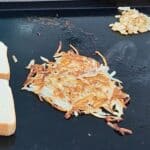 Golden brown shredded hash browns cooking on a Blackstone griddle, with diced onions sautéing nearby and slices of bread to the side.