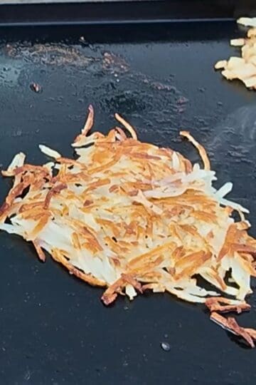 Golden brown shredded hash browns cooking on a Blackstone griddle, with diced onions sautéing nearby and slices of bread to the side.