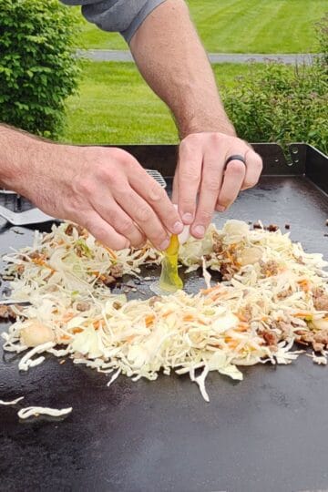 Egg roll in a bowl cooking on a Blackstone griddle with an egg cracked into the center of cabbage, carrots, and ground beef.