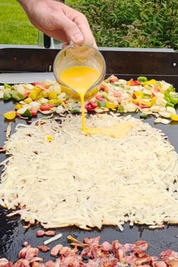 Hashbowns, peppers, onions, and bacon on a Blackstone Griddle. Eggs pouring on the hasbrowns.