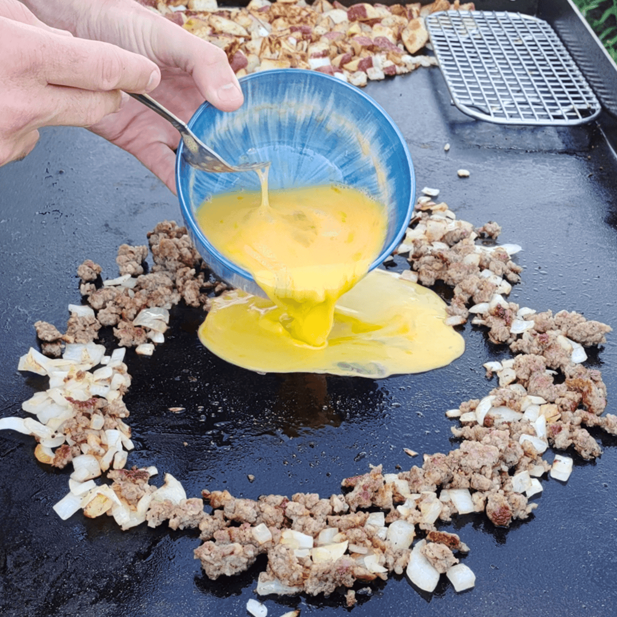 Scrambled eggs being poured into the center of a sausage and onion ring on a Blackstone griddle, with diced potatoes cooking in the background.