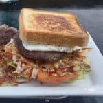 A hearty breakfast sandwich on toasted Texas toast, stacked with a fried egg, thick sausage patty, crispy hash browns, grilled onions, tomato slices, and lettuce, served on a white plate atop a flat top griddle.
