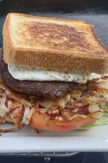 A hearty breakfast sandwich on toasted Texas toast, stacked with a fried egg, thick sausage patty, crispy hash browns, grilled onions, tomato slices, and lettuce, served on a white plate atop a flat top griddle.