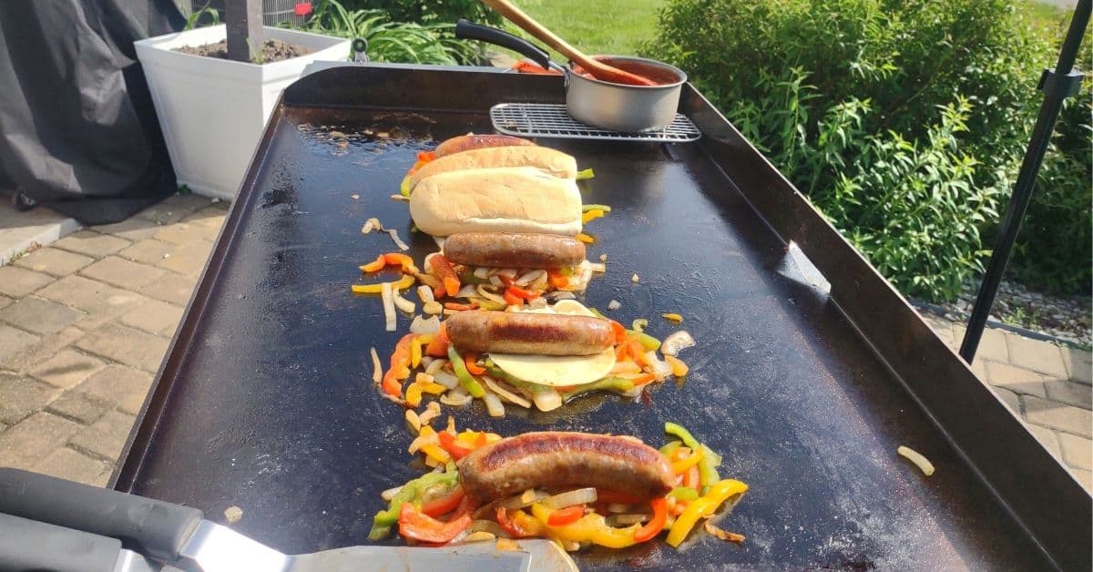 Italian sausages on peppers on a blackstone griddle with a bun on top of one of them.