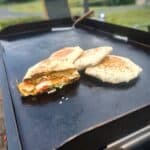 Three grilled crunchwraps cooking on a Blackstone griddle, with one sliced in half to show layers of seasoned ground beef, lettuce, tomato, cheese, and a crispy tostada shell inside a toasted tortilla. The griddle is outdoors with a spatula in the background.