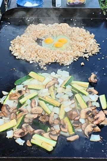 Vegetables and fried rice cooking on a flat-top griddle. Zucchini, mushrooms, and onions are sautéing in the front, while a mound of rice surrounds three cracked eggs in the back, ready to be mixed for hibachi-style fried rice.