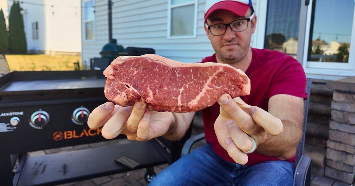 Man holding a raw, marbled steak in front of a Blackstone griddle outdoors.