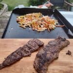 Grilled skirt steak resting on a cutting board with peppers and onions cooking on a Blackstone griddle in the background.